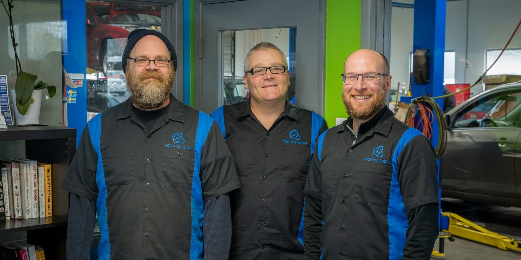 Travis Decker, owner, in the middle, with two of the Atomic Auto crew in the shop's blue and black work shirts, a car up on a lift through the window behind them
