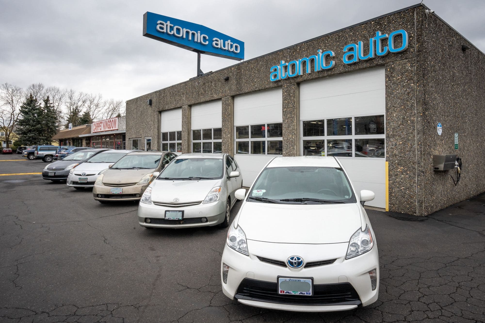 Priuses parked in a row outside Atomic Auto's four bays, the blue atomic auto sign above them under a grey sky
