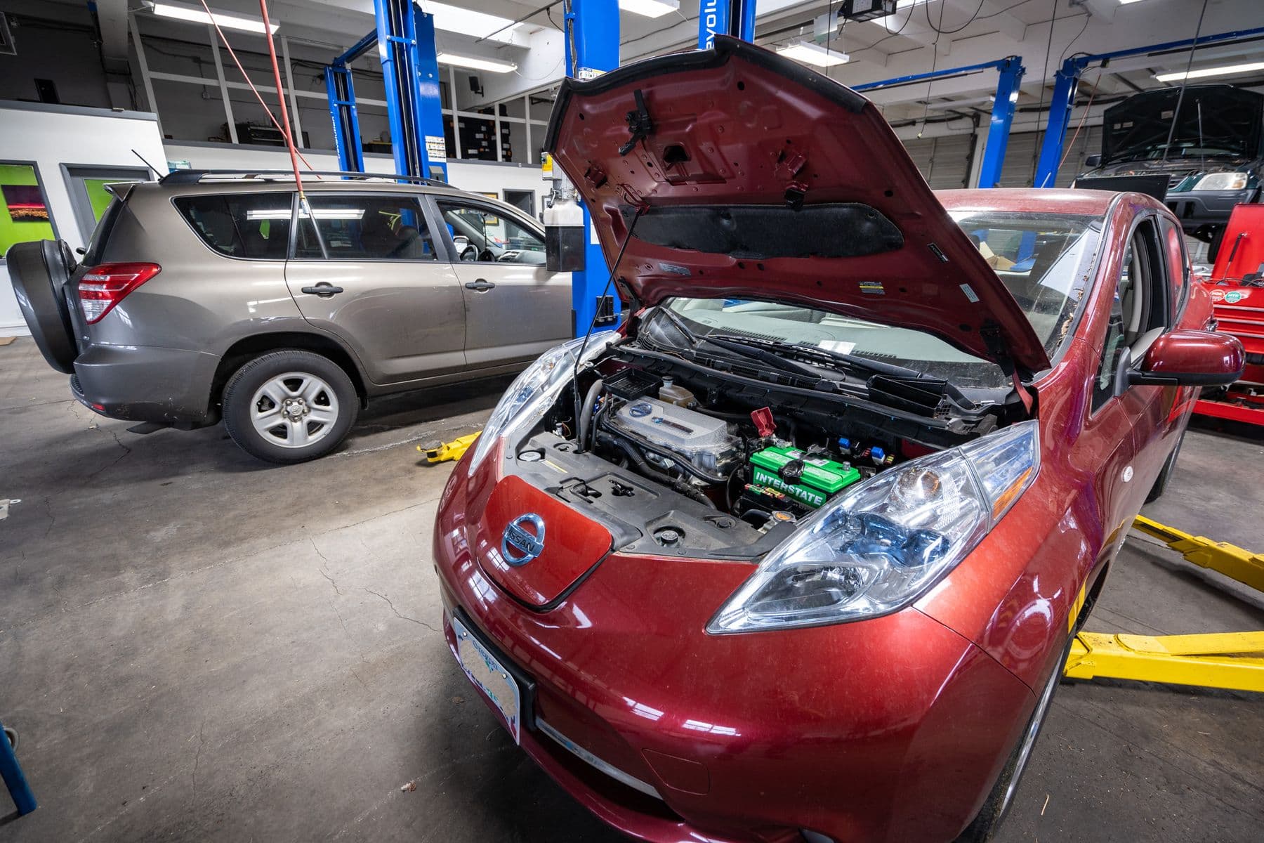 A red Nissan Leaf with its hood up on the lift at Atomic Auto, a RAV4 behind it